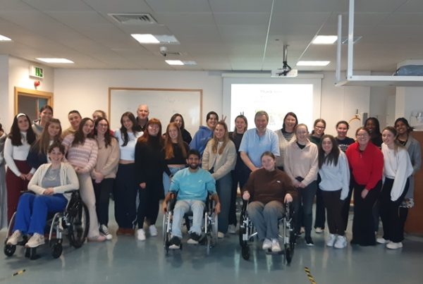 A group of diverse people, including individuals in wheelchairs, pose together in a room with a projector screen and whiteboard in the background. They appear to be in a friendly and inclusive setting at Trinity College Dublin, with smiles and casual attire.